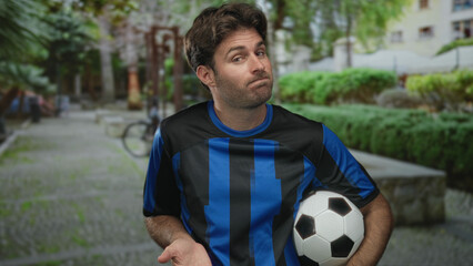 Man holding soccer ball shrugs shoulders in park wearing blue striped jersey and casual sportswear; team support disappointment.