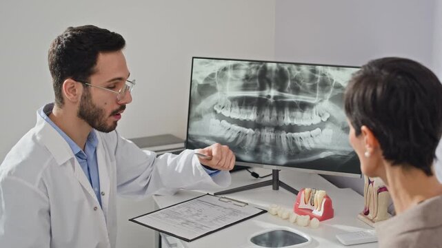 Waist up of Middle Eastern male dentist showing patient dental x-ray and demonstrating treatment options with prosthetic tooth model in clinical office setting