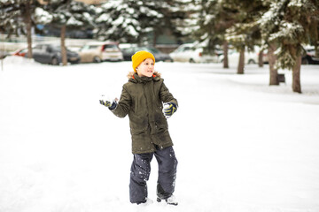 A cheerful child in a yellow beanie and green winter coat plays with snowballs in a snowy park surrounded by evergreen trees.