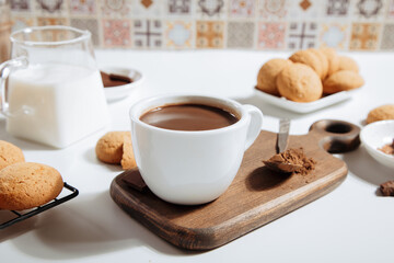 Сup of hot chocolate on a wooden tray surrounded by cookies, milk, and cocoa powder on a white table.