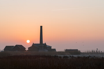 A sunrise over a field with a large brick building in the background. The sky is orange and the sun is rising.