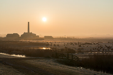 A sunrise over a field with a large brick building in the background. The sky is orange and the sun is rising. In the front you see the pasture with a wooden fence.