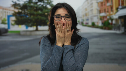 Middle aged woman with black glasses covers her mouth with both hands on street in a city square  happiness. © Krakenimages.com