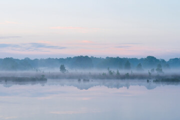 In the early morning a lake with a reflection of the sky and the trees. The sky is overcast and the air is cool and damp. The scene is quiet and peaceful, with the lake providing a sense of solitude a