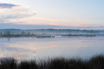 Obraz premium In the early morning a lake with a reflection of the sky and the trees. The sky is overcast and the air is cool and damp. The scene is quiet and peaceful, with the lake providing a sense of solitude a