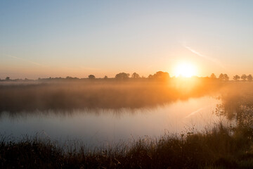 Obraz premium A sunrise over a lake in the Netherlands. The sky is orange and the sun is rising at the horizon.
