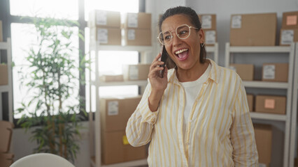 Woman holds smartphone to ear and smiles amid cardboard boxes on shelving in a building; small...