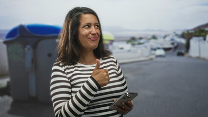 Woman brunette middleage hispanic holding smartphone with hands visible, smiling while looking at screen on street near trashcan and cars; everyday connection joy.