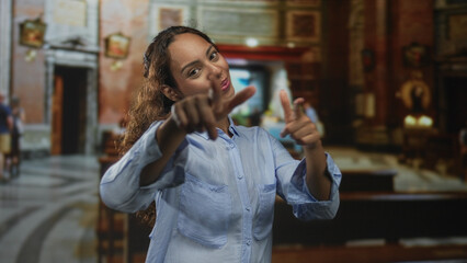 Woman points finger to camera in church building while smiling and gesturing with both hands; joyful welcome.