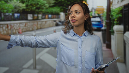 Young african american woman hailing a taxi on a street, holding a clipboard and pointing with her right arm; urban survey confidence.