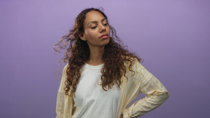 Fototapeta premium Woman with hand on hip and curly hair wearing white t shirt and yellow striped overshirt in studio with purple backdrop; confidence reflection.