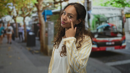 Fototapeta premium Woman points finger to cheek on street near a bus and trees, wearing striped shirt and white tee; urban confidence.
