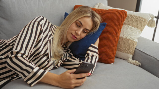 Woman lying on comfortable striped couch with pillow resting head scrolling smartphone in building; relaxation calm.