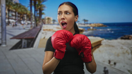 Hispanic woman wearing red boxing gloves holds fists in guard on a seaside street promenade; determination training.