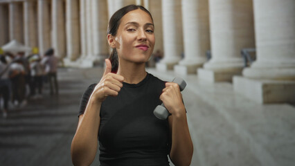 Young hispanic woman holding a dumbbell and giving thumbs up beside old stone building colonnade in town; confidence wellness.