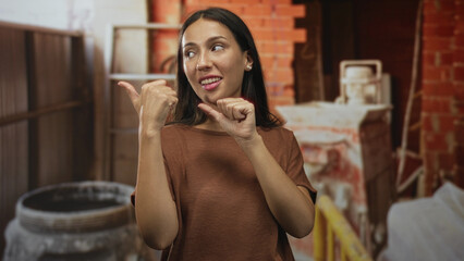 Hispanic woman with thumbs pointing to self in building construction site indoors wearing a brown shirt; confidence.