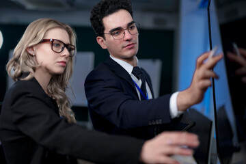 A male team leader in a suit uses a stylus to explain a complex project on a large screen, providing one-on-one training to a female colleague in a modern, high-tech office.
