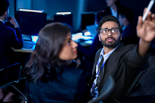 A diverse team of professionals collaborates on a business strategy late at night. A male manager uses a tablet and stylus to explain key points to his female colleague in a modern boardroom.