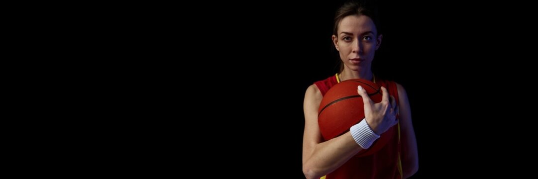 Female athlete holding basketball with intense gaze against dark background. Concept of focus, determination, and inner power for training and sports promotion. Banner - Powered by Adobe