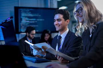 A young male professional in a suit smiles as he points out a successful result on a computer screen to his female colleague, celebrating a project milestone in a modern tech office.