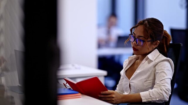 A diligent business woman in glasses works attentively at her desk, taking notes in a red notebook. She is focused in a modern office environment with coworkers nearby.
