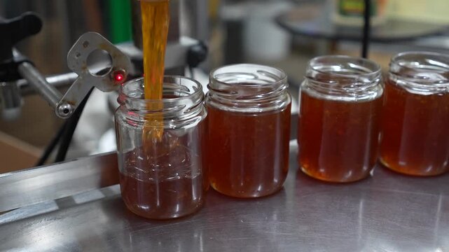 Liquid honey is being poured into a glass jar on a production line, showcasing the filling process with a close-up view, emphasizing the smooth flow and texture