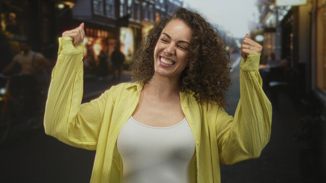 Young woman with curly hair raises fists in a city street wearing yellow shirt and white tanktop smiling confidently amid evening storefronts; joy celebration.
