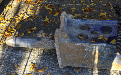 Heavy-duty blue and rusty hydraulic breaker (rock hammer) attachment for an excavator, lying on sunlit asphalt scattered with brown and yellow autumn leaves.