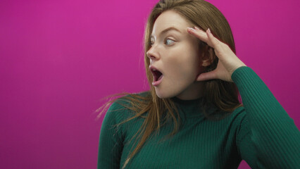 Woman in green sweater cups her hand to ear and opens mouth in pink studio with curious tilt of...