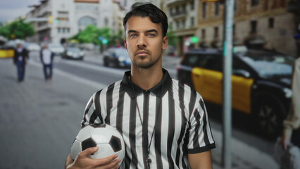 Young hispanic man in referee uniform holding soccer ball standing on city street with blurred urban background, highlighting the dynamic outdoor environment.