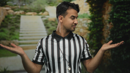 Young hispanic male referee outdoors gesturing with hands extended to each side, wearing black and white striped shirt, conveying choice or decision making in a serene garden setting.