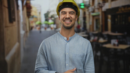Fototapeta premium Young hispanic man with hardhat laughs on outdoor restaurant terrace, conveying joy and industry in a vibrant café street setting, blending work and leisure.