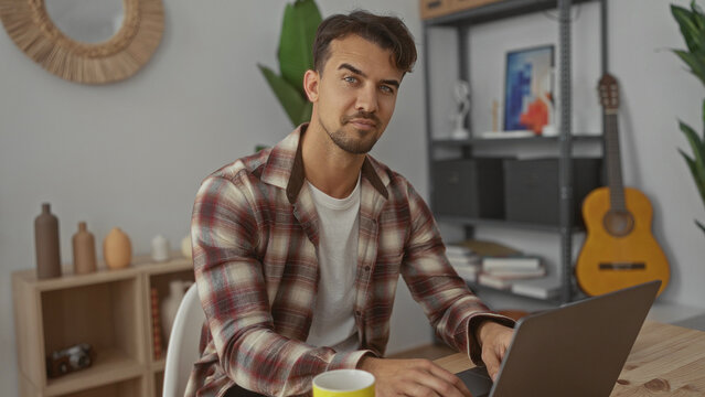 Young hispanic man working on laptop in modern office interior with guitar visible in background showing thoughtful expressions