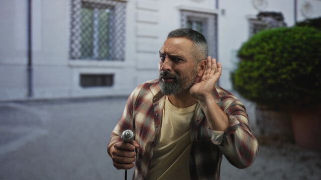 Man holding a power drill and cupping his ear by a building facade, leaning forward with a pained listening expression; safety concern. - Powered by Adobe