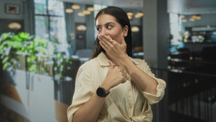Young brunette woman covering mouth with hand and thumb pointing to herself in restaurant building; surprise playful moment.