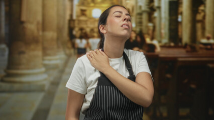 Obraz premium Young brunette woman in pinstripe apron holding hand to chest for heartattack inside church building while wincing in discomfort; faith struggle pain.