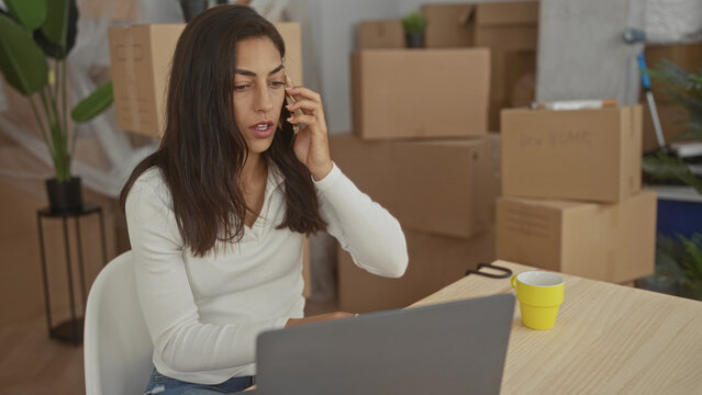 Woman holds phone to ear while typing on laptop amid packed moving boxes in building; moving stress.