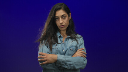 Woman with arms crossed over chest wearing denim shirt looking down in deep blue studio; rejection...