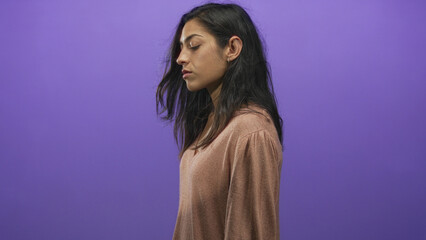 Woman in studio with face in profile and closed eyes against a purple backdrop, wearing brown v neck top and small hoop earring; contemplation calm.