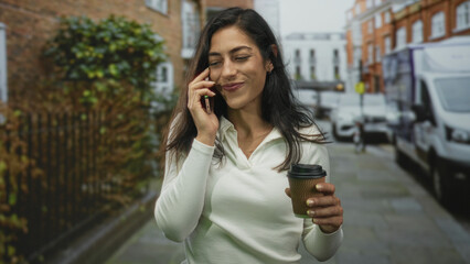 Woman talking on phone holding coffee cup while walking briskly down a city street past brick building and delivery van; morning commute joy.