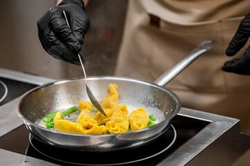 Close-up of a professional chef wearing black gloves, using a spoon to stir fresh, handmade tortellini pasta with green peas in a stainless steel frying pan with a creamy white sauce in a modern kitch