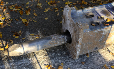 Detailed view of the worn, pointed steel chisel bit of a heavy-duty hydraulic breaker attachment, resting on dark asphalt scattered with brown and yellow autumn leaves.