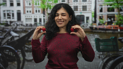 Woman young brunette holding two strawberries with bare hands and smiling on a street in amsterdam; playful joy.