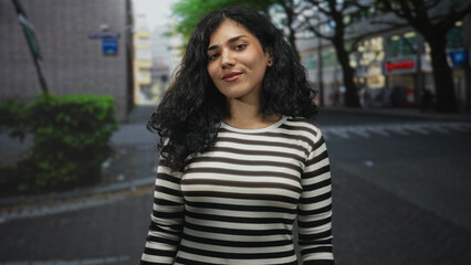 Young arab woman smiling with cheek piercings and striped top standing on a city street near trees and shops; confidence.
