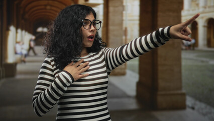 Young arab woman wearing striped shirt and glasses points finger with hand on chest in an old building corridor; surprise curiosity.