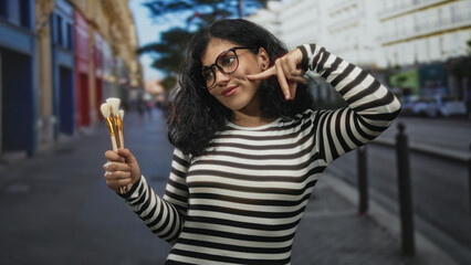 Fototapeta premium Young arab woman wearing glasses and striped top holds brushes and points finger to cheek on a street in a city scene; creativity playful expression.