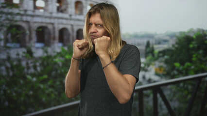 Blond man with fists raised in fighting stance at roman coliseum ruins balcony overlooking...