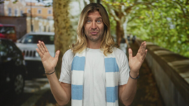 Man wearing striped soccer scarf raises hands on street lined with parked cars and autumn trees; team spirit passion. - Powered by Adobe