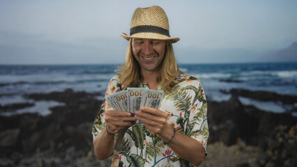Blond man in straw hat counting hundred dollar bills with hands on street; vacation financial freedom joy.