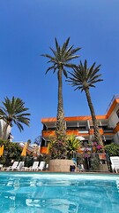 Palm trees and modern hotel with orange balconies by swimming pool, blue sky, sunny day, summer vacation